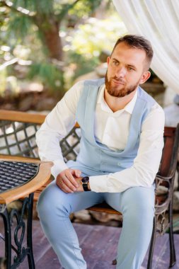 Mavi takım elbiseli bir damat parkta bir sandalyede oturuyor.. 