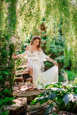 Uzun bukleli güzel bir gelin parktaki bir bankta otların ve ağaçların arasında oturur..