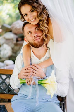  Uzun bukleli gelin nazikçe omuzlarına sarılıyor mavi takım elbiseli damat.