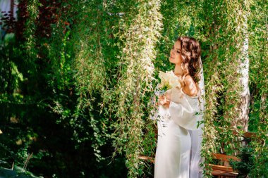 Kıvırcık elbiseli, omuzları açık gelin ve söğüt ağacında bir buket çiçek.