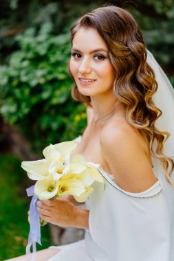 Uzun bukleli güzel bir gelin parktaki bir bankta otların ve ağaçların arasında oturur..