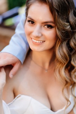 Damat uzun bukleli gelini nazikçe kucaklıyor. Düğün fotoğrafı çekimi yaz bahçesinde.