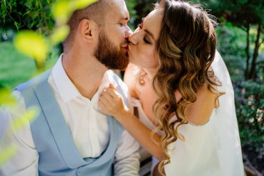 Mutlu ve güzel gelin ve damat parkta nazikçe öpüşürler. Bahçedeki fotoğraf çekiminde aşık bir çift..