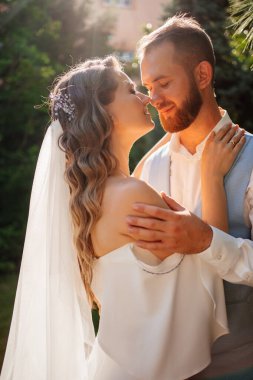 Güneş parlaması. Yaz bahçesinde yürüyen yeni evliler. Mavi takım elbiseli, uzun kıvırcık elbiseli, omuzları açık beyaz zambak buketli güzel bir damat. Park 'ta düğün fotoğrafı çekimi.