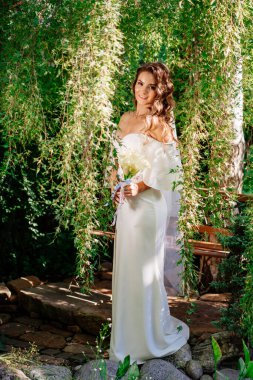 Uzun bukleli gelin, açık omuzlu beyaz bir elbise ve söğüt ağacında bir buket zambak duruyor. Parkta düğün fotoğrafı çekimi var..