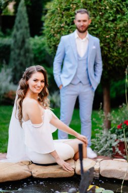 Yeni evliler, bir yaz parkındaki yapay bir göletin yanında. Mavi takım elbiseli güzel damat ve zarif beyaz elbiseli bukleli gelin. düğün fotoğrafı çekimi.