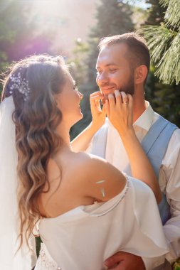 Güneş parlaması. Yaz bahçesinde yürüyen yeni evliler. Mavi takım elbiseli, uzun kıvırcık elbiseli, omuzları açık beyaz zambak buketli güzel bir damat. Park 'ta düğün fotoğrafı çekimi.