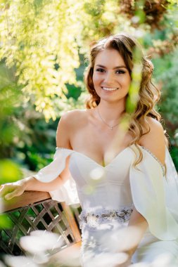 Uzun bukleli, uzun elbiseli güzel bir gelin parktaki bir bankta çimenlerin ve ağaçların arasında oturur. düğün fotoğrafı çekimi.