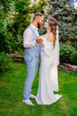 Yaz bahçesinde yürüyen yeni evliler. Mavi takım elbiseli güzel bir damat ve geniş omuzlu uzun bukleli bir gelinlik ve bir buket beyaz zambak. Parkta düğün fotoğrafı çekimi var..
