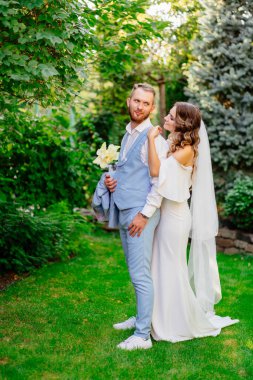 Yaz bahçesinde yürüyen yeni evliler. Mavi takım elbiseli güzel bir damat ve geniş omuzlu uzun bukleli bir gelinlik ve bir buket beyaz zambak. Parkta düğün fotoğrafı çekimi var..