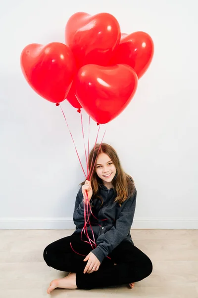 身穿深色衣服、心形气球的少女靠着白墙坐着. — 图库照片