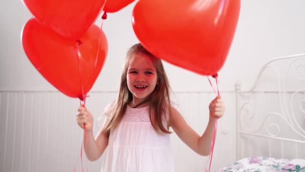 Rolig flicka med röda ballonger i form av hjärtan ler på sängen av vit vägg — Stockvideo