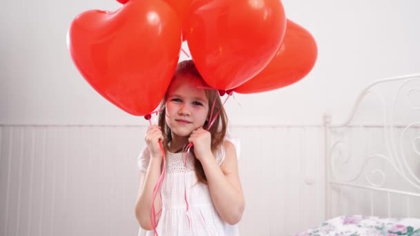 Menina engraçada com balões vermelhos em forma de corações sorrindo na cama pela parede branca — Vídeo de Stock