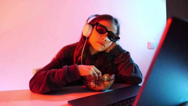 Menina em óculos para 3D e fones de ouvido assiste filme triste no laptop, come pipoca — Vídeo de Stock