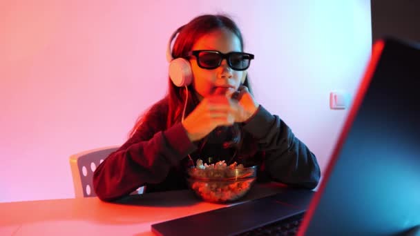 Menina em óculos 3D e fones de ouvido assiste filme terrível no laptop, come pipoca — Vídeo de Stock