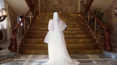 Güzel, uzun gelinlikli, trenli gelin altın merdivenlerden tırmanıyor..