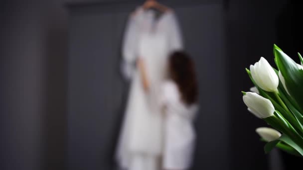 Bride in a silk robe next to a white wedding dress hanging on the closet. — Stock Video