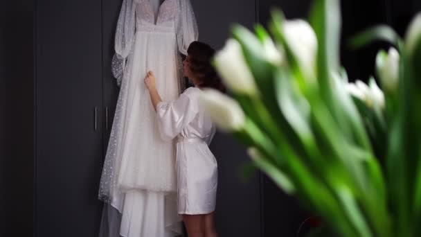 Novia en una bata de seda junto a un vestido de novia blanco colgando en el armario. — Vídeos de Stock
