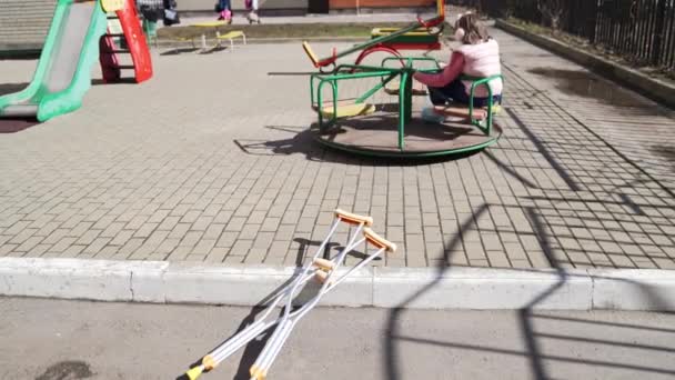 Teenie-Mädchen mit gebrochenem Bein auf Krücken sitzt auf einem Karussell auf dem Spielplatz. — Stockvideo
