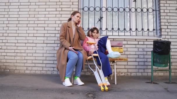 Mom talking on phone, daughter with crutches on bench in yard — Stock Video