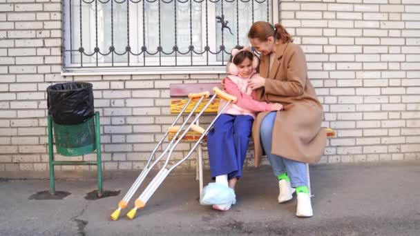 松葉杖を持つ娘の母親がアパートの庭のベンチに座り — ストック動画