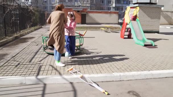 Mama pomaga córce ze złamaną nogą stanąć na kulach na placu zabaw. — Wideo stockowe
