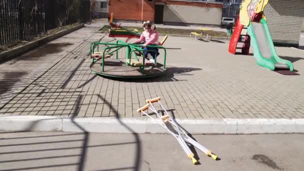 Teen girl with a broken leg on crutches sits on a carousel in the playground. — Stock Video