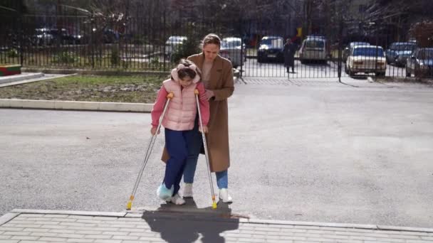 Moeder helpt haar dochter om op krukken te leren lopen. gebroken helling en een hoge stoeprand. — Stockvideo