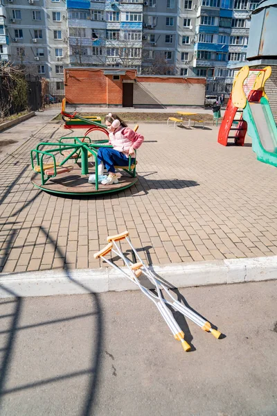 Menina adolescente com uma perna quebrada em muletas senta-se em um carrossel no parque infantil. — Fotografia de Stock