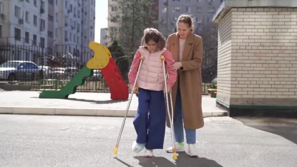 Mom helps daughter learn to walk on crutches. plaster on your feet. — Stock Video