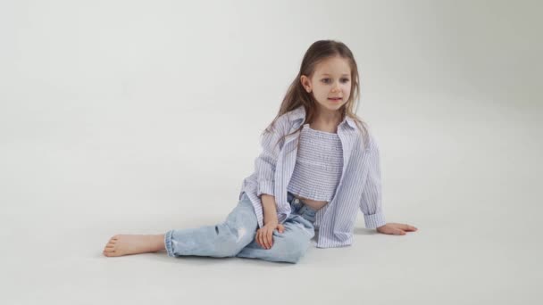 Bambina con i capelli lunghi in jeans e camicia posa su uno sfondo bianco. — Video Stock