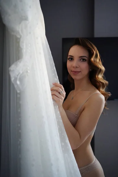 Nevěsta s dlouhými vlasy ve spodním prádle vedle bílých svatebních šatů — Stock fotografie