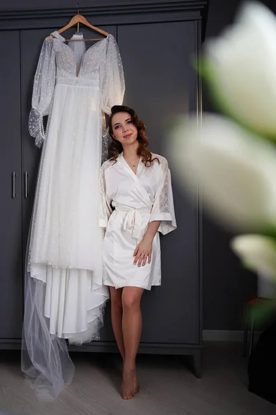 Novia en una bata de seda junto a un vestido de novia blanco colgando en el armario. — Foto de Stock