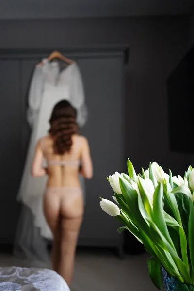 Noiva em roupa interior e meias ao lado de vestido de noiva branco pendurado no armário. — Fotografia de Stock