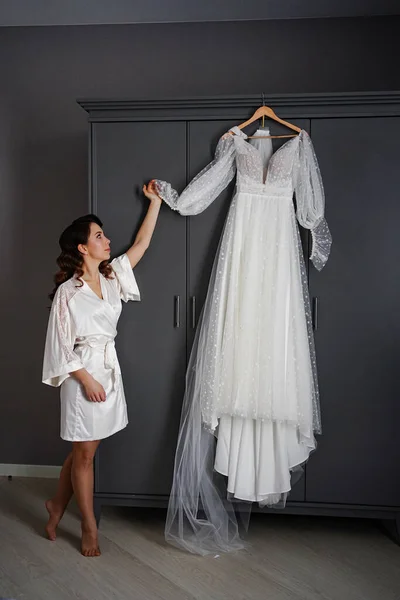 Noiva em um manto de seda ao lado de um vestido de noiva branco pendurado no armário. — Fotografia de Stock