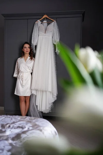 Bruid in een zijden mantel naast een witte trouwjurk hangend aan de kast. — Stockfoto