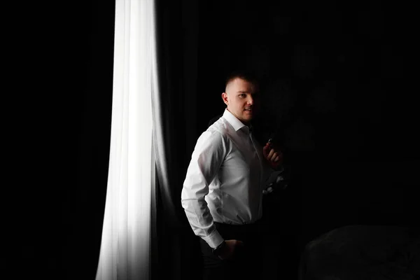 Lächelnder Mann in weißem Hemd mit Jacke in der Hand am Fenster in einem dunklen Raum — Stockfoto