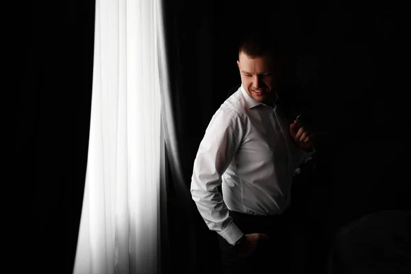 一个穿着白衬衫，手里拿着一件夹克的男人在一间黑暗的房间的窗边. — 图库照片