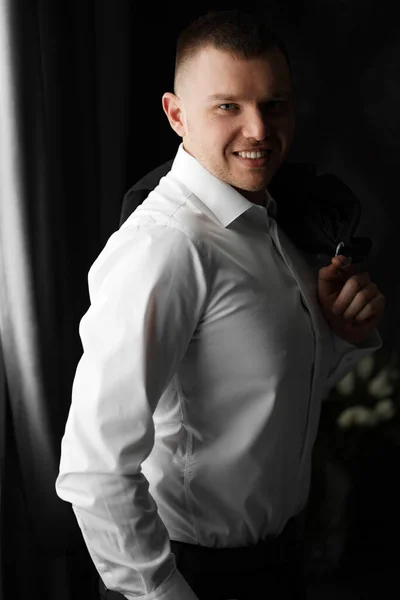 Χαμογελαστός άντρας με λευκό πουκάμισο με σακάκι στα χέρια δίπλα στο παράθυρο σε σκοτεινό δωμάτιο — Φωτογραφία Αρχείου