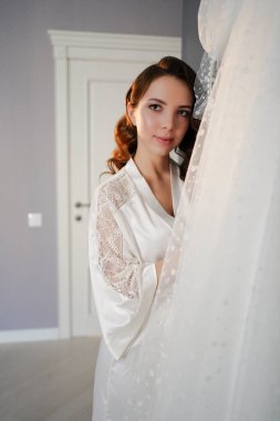 Uzun saçlı, ipek bornozlu gelin, beyaz gelinliğin yanında.