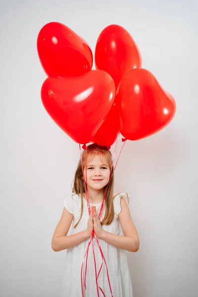 Funny girl with red balloons smiling,folds palms in front of near him white wall