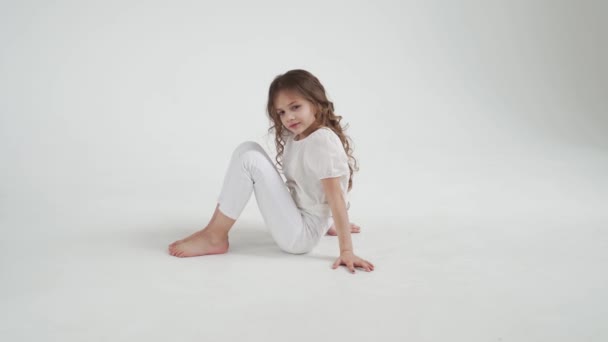 Piękna dziewczynka z długimi włosami w białych ubraniach siedzi na podłodze. — Wideo stockowe