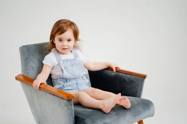 En vacker liten flicka i denim overall sitter i en stol. — Stockfoto