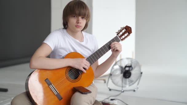 Young guy in a white T-shirt learns to play guitar. online lessons on smartphone — Stock Video