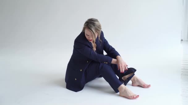Mulher de terno azul senta-se descalço no estúdio de fotos branco e segura sapatos de salto — Vídeo de Stock