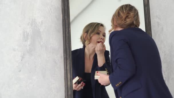 Una hermosa mujer rubia en un traje azul hace maquillaje de labios de pie junto al espejo. — Vídeo de stock