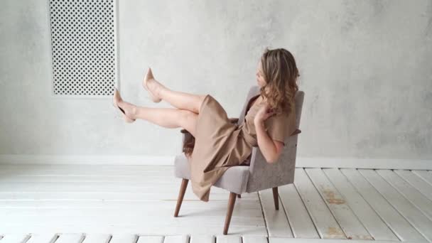 Eine blonde Frau im beigen Kleid posiert auf einem grauen Stuhl in einem Fotostudio. — Stockvideo