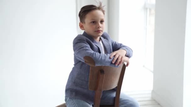 Хлопчик-підліток в піджаку і джинси позує на стілець біля вікна . — стокове відео