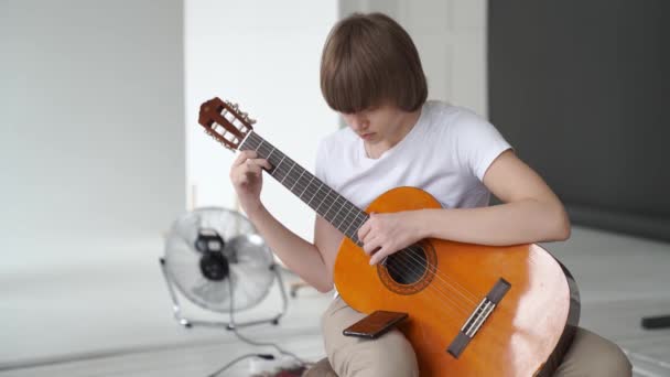 Un joven con una camiseta blanca aprende a tocar la guitarra. — Vídeo de stock