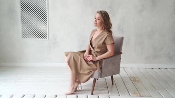 Uma mulher loira em um vestido bege posa em uma cadeira cinza em um estúdio de fotos. — Vídeo de Stock